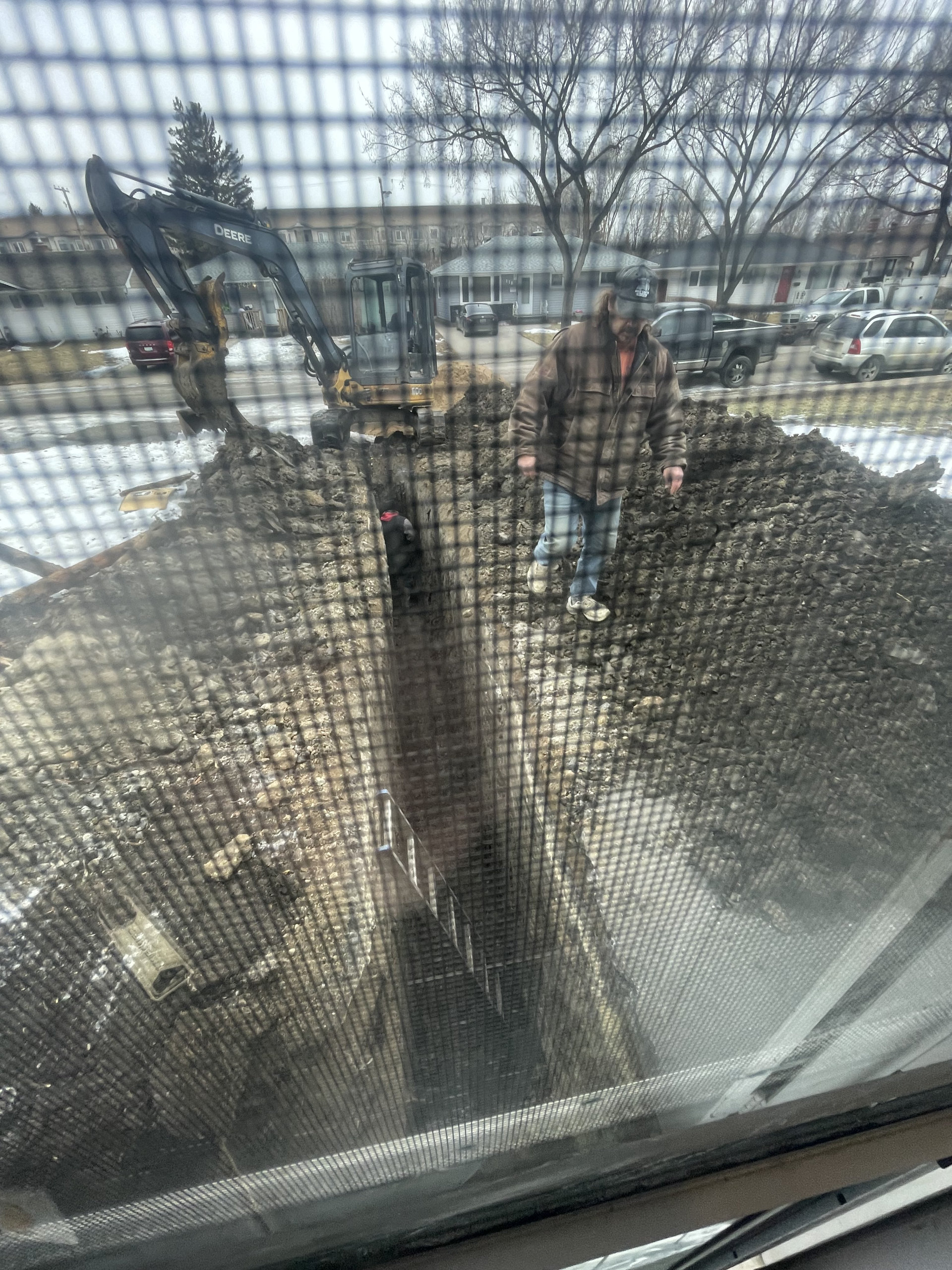 A construction worker stands next to a deep trench prepared for sewer root removal, with a backhoe nearby, viewed through a window screen on a cloudy day.